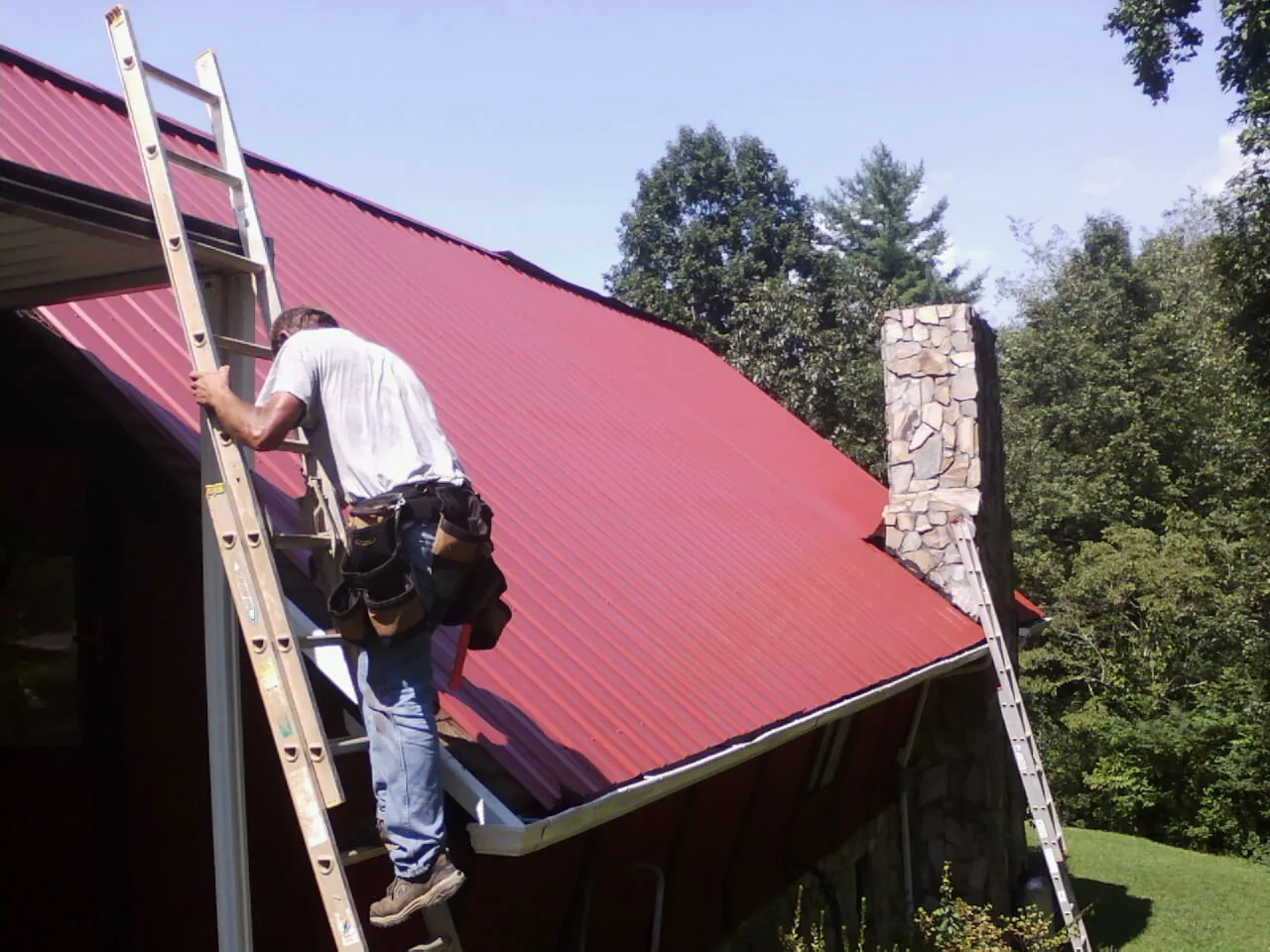 Roofer on a ladder beside a red metal roof and stone chimney — Soffit & Fascia Repair by Roofing Brothers in Appalachia